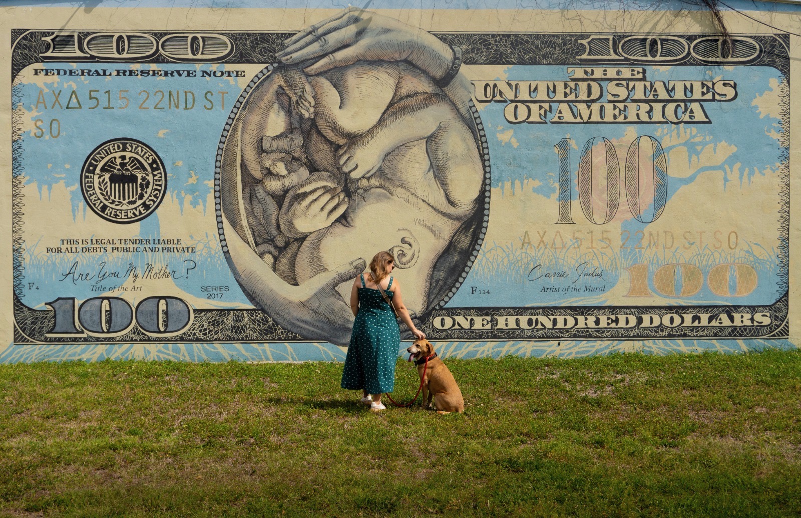 A woman with her dog standing in front of a giant hundred-dollar-bill mural — the everyday weight of money.